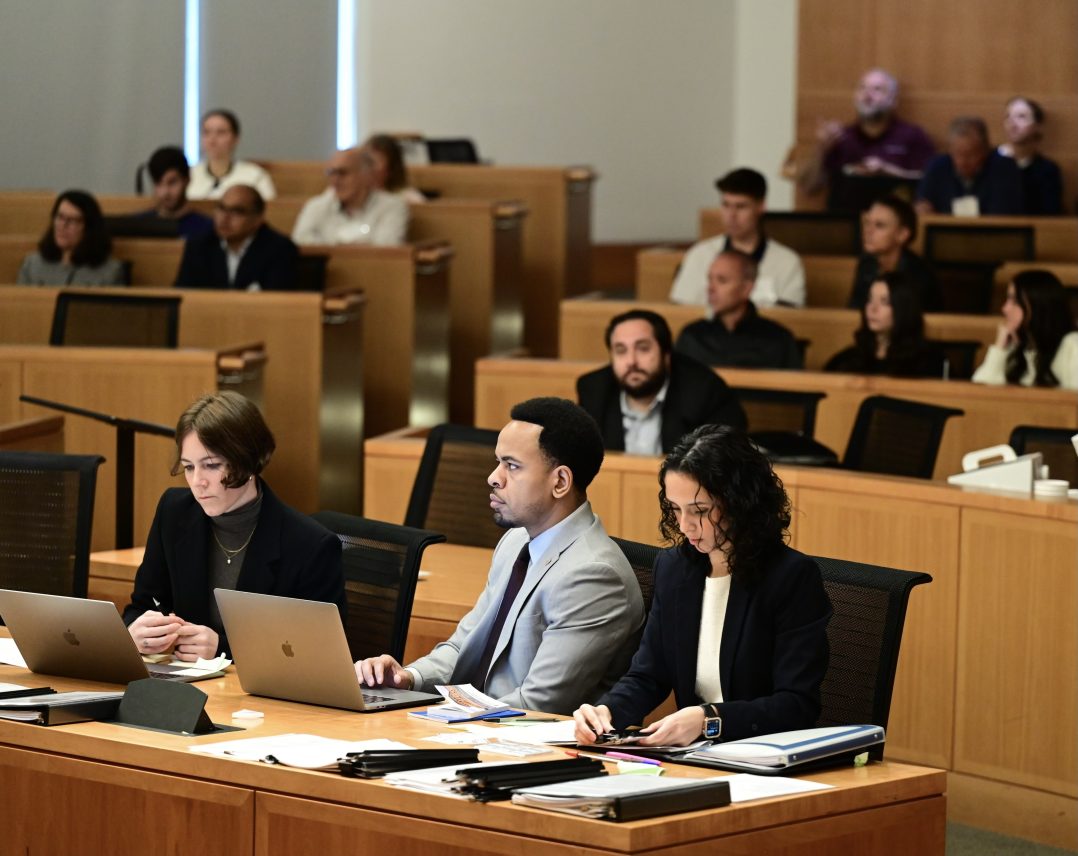 Attendees listening attentively in a moot courtroom setting, some using laptops, during a mock trial case.